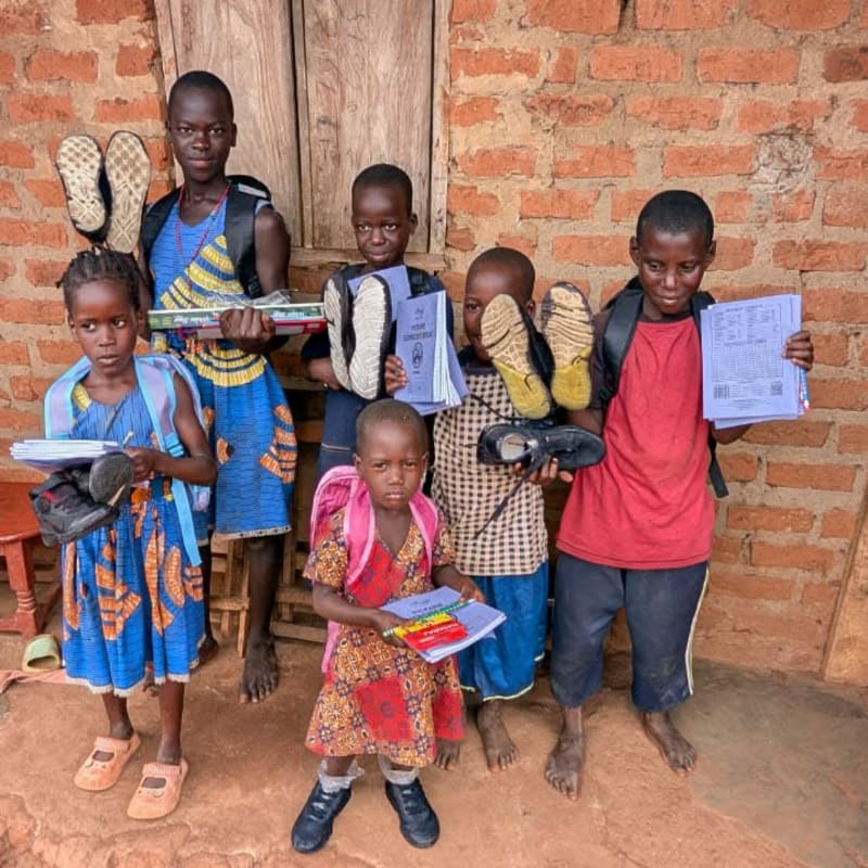 Ugandan children holding school books, shoes, and backpacks outside a brick home.