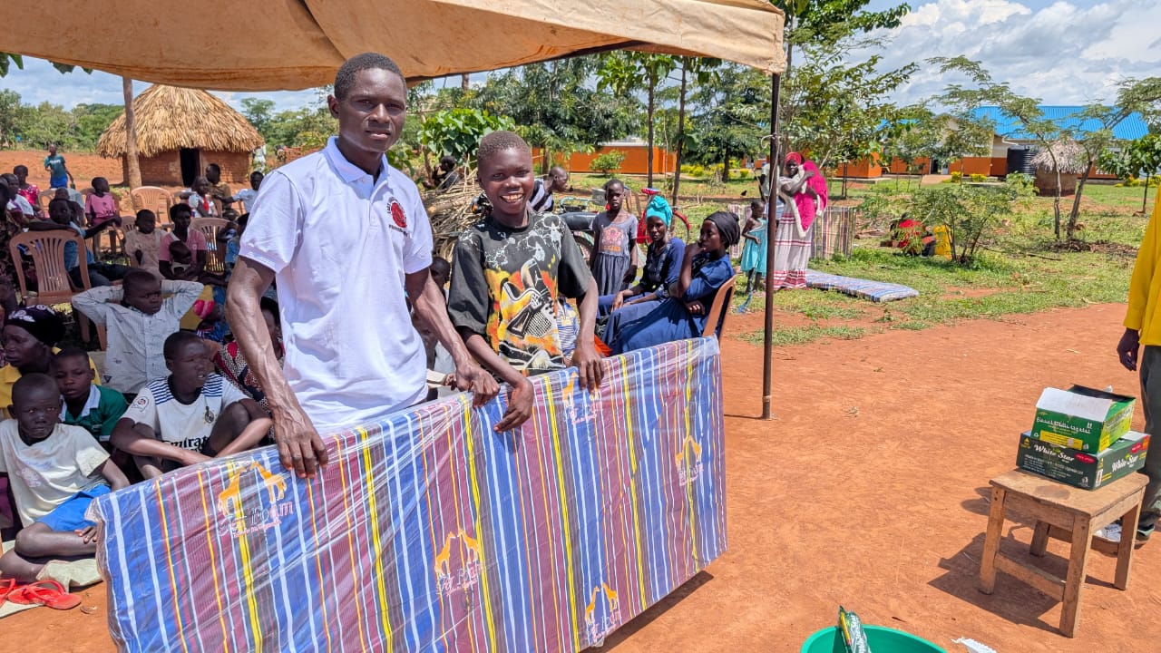 A recipient smiling while receiving a mattress in front of the gathered community.