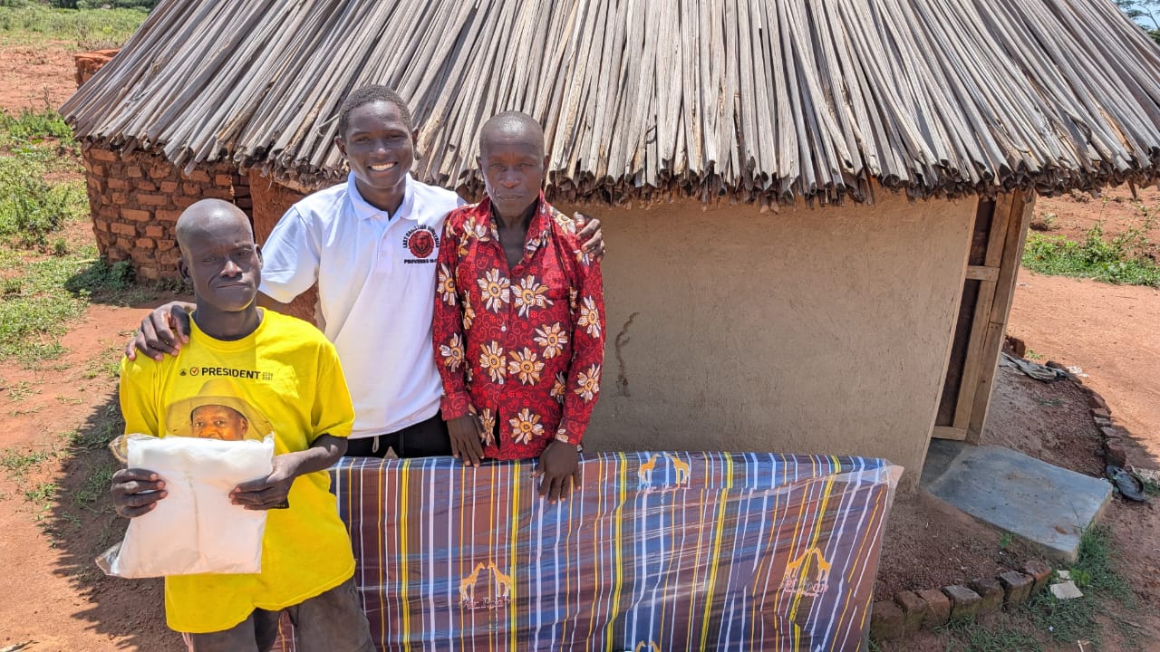 Two recipients standing with a mattress and food assistance outside a home in Uganda.