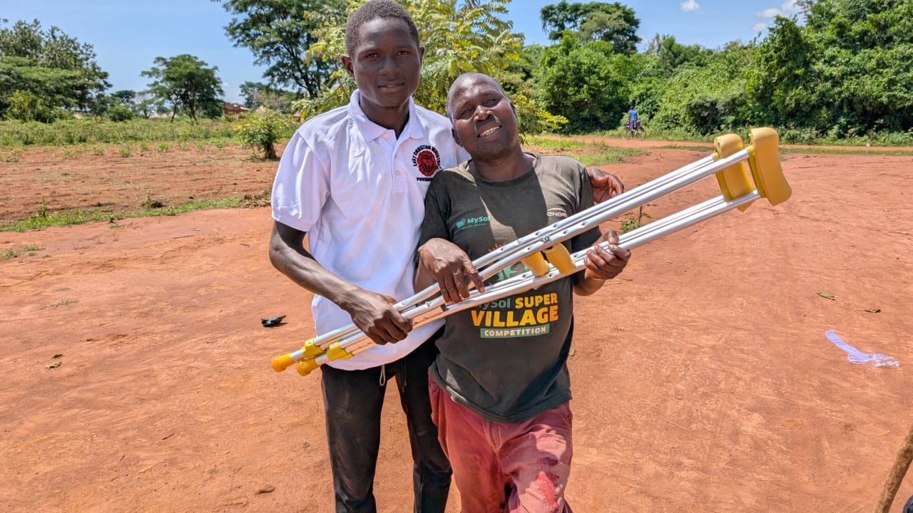 An older man holding new crutches beside Kutesa Henry outdoors in Uganda.