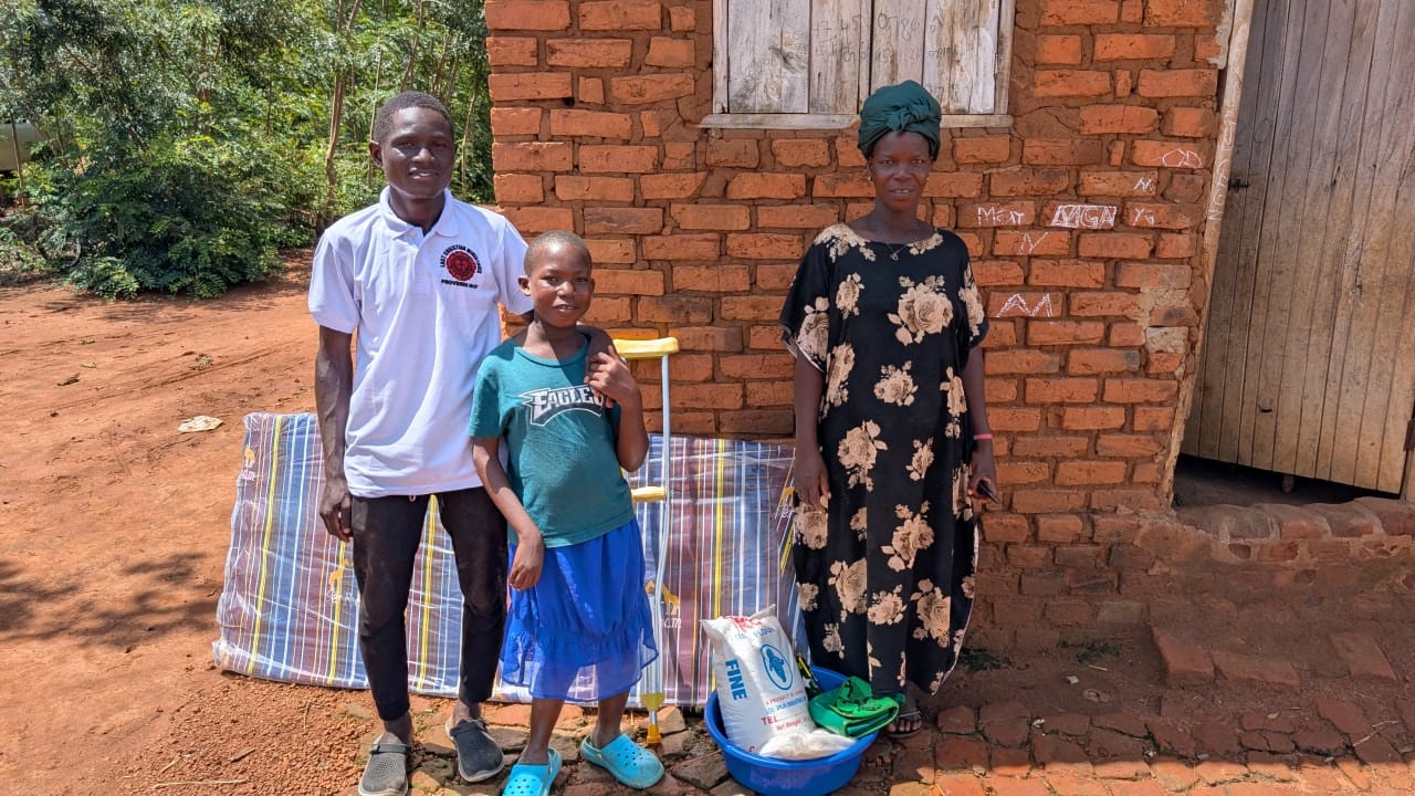 A family in Uganda standing outside a brick home with a mattress, food, basin, and crutch support provided through the Easter distribution.