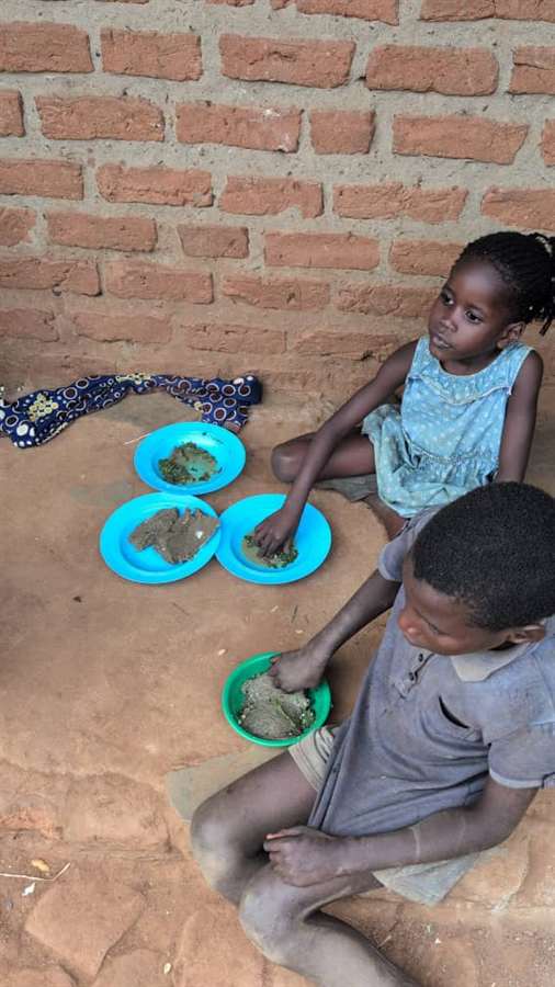 A small child eating from a bowl while another plate of dirt and greens sits nearby, showing severe food deprivation.