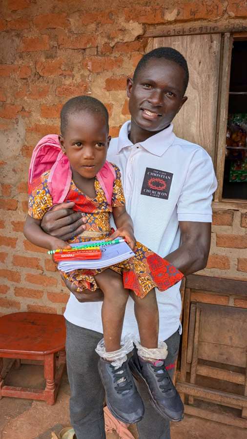 Kutesa Henry holding a small child and school supplies outside a brick home in Uganda.